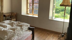 Une chambre avec un plafond en bois, un lit recouvert de linge de lit blanc, deux fenêtres donnant sur la verdure et une lampe.