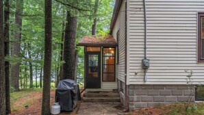 Une cabane avec une entrée en pierre, entourée de grands arbres, et un objet couvert situé près de la porte.