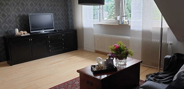 Un soggiorno moderno con un mobile TV nero, una TV a schermo piatto, un tavolino in legno con oggetti decorativi, un tappeto rosso e una finestra vista verde.