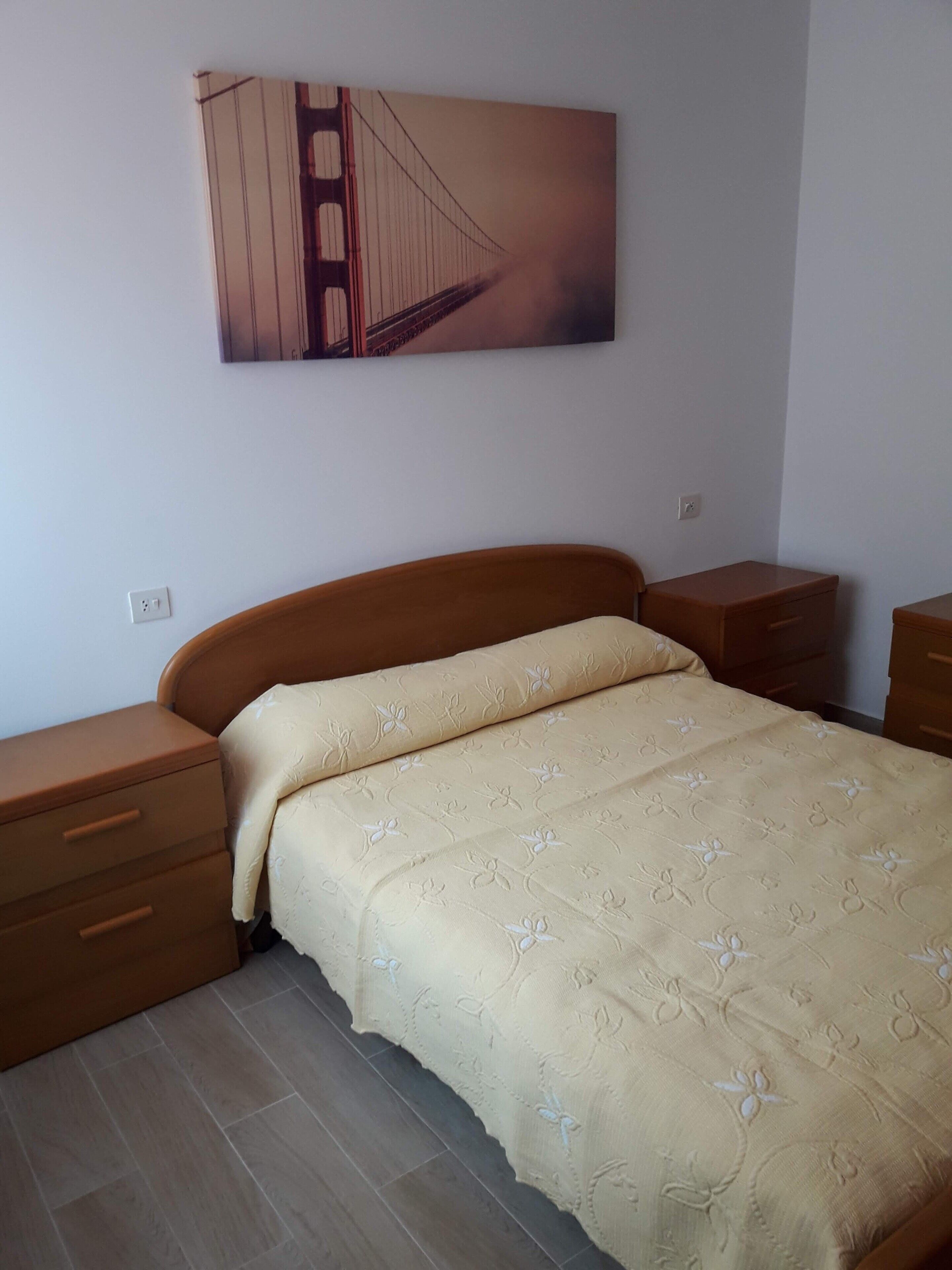 Ein Schlafzimmer mit einem Bett, zwei Nachttischen, einem an der Wand hängenden Bild einer Brücke und einer Steckdose.