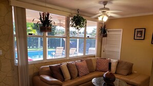 A living room with a brown leather sofa, a glass coffee table, and a ceiling fan. There are windows with curtains, a view of a pool, and outdoor furniture.