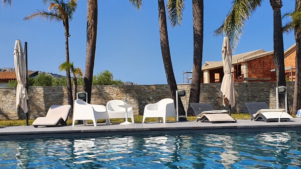Un área de piscina con sillones blancos y sombrillas, rodeada de palmeras y un muro de piedra.