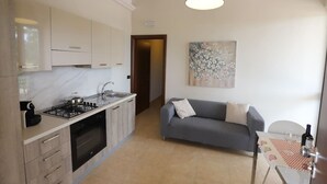 Cocina compacta con estufa a gas, horno y armarios, un sofá gris y una mesa con una botella de vino.