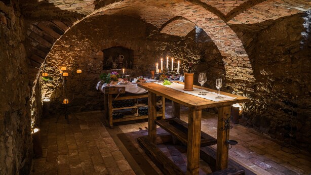A rustic cellar with a wooden table set for dining, surrounded by stone walls and brick arches.