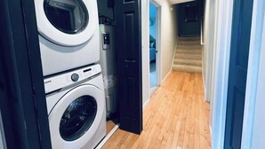 Un lavadero con lavadoras y secadoras apiladas, un calentador de agua y una puerta que da a un pasillo con una escalera.