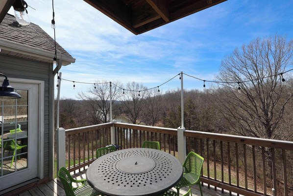 A covered outdoor deck with a round table and chairs, string lights, and a view of trees and hills.