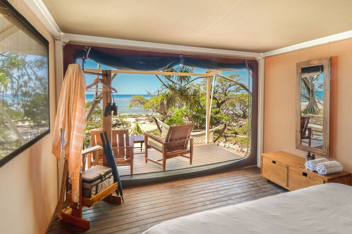 Una habitación con piso de madera, una cama, un banco de madera, una silla y vista a la playa y al océano.