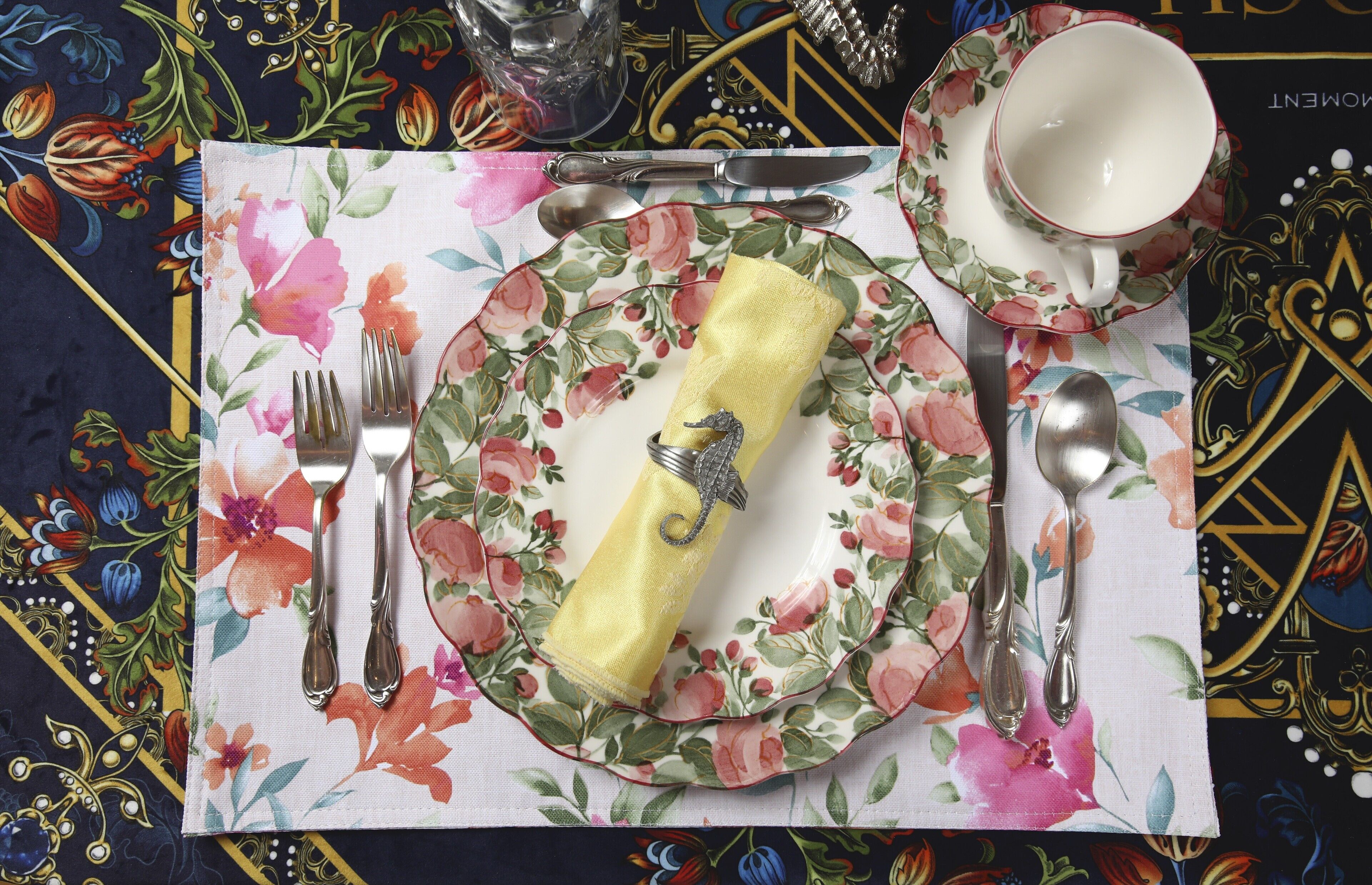 Une table dressée avec une nappe à motifs floraux, un porte-serviette, des couverts et une tasse avec sa soucoupe.