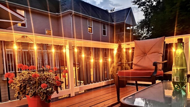 A wooden deck with string lights, a potted plant with red flowers, and a glass table. - Charlotte's Rose Inn (Charlottetown)