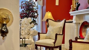 A traditional living room with a red accent wall, a white chair with a decorative pillow, a glass coffee table, and a large white orchid plant. - Charlotte's Rose Inn (Charlottetown)