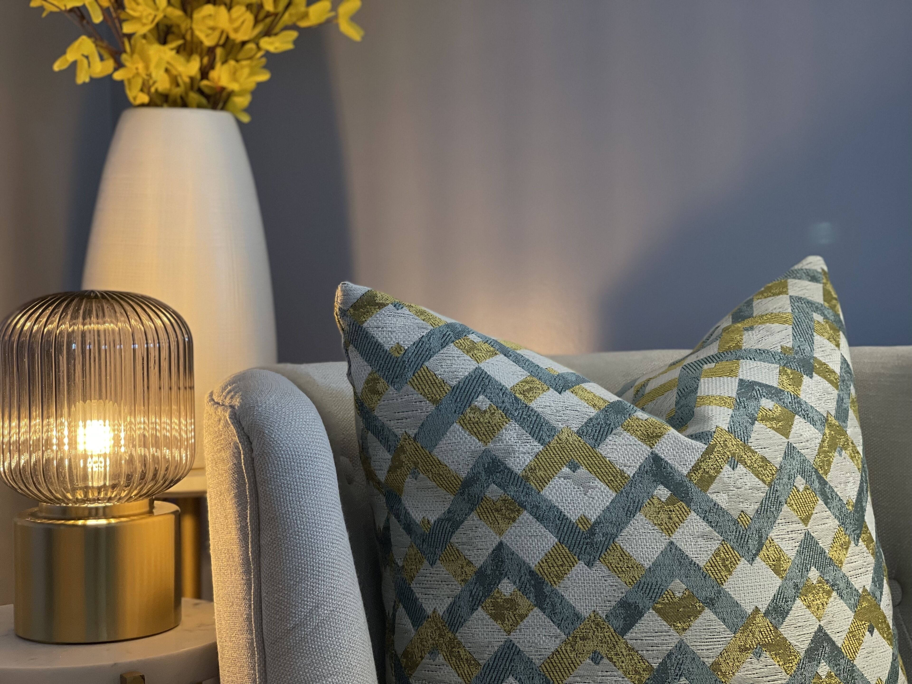 Um candeeiro com base canelada, uma almofada com padrão de losangos e um vaso com flores amarelas sobre uma mesa branca.