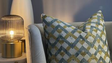 Um candeeiro com base canelada, uma almofada com padrão de losangos e um vaso com flores amarelas sobre uma mesa branca.