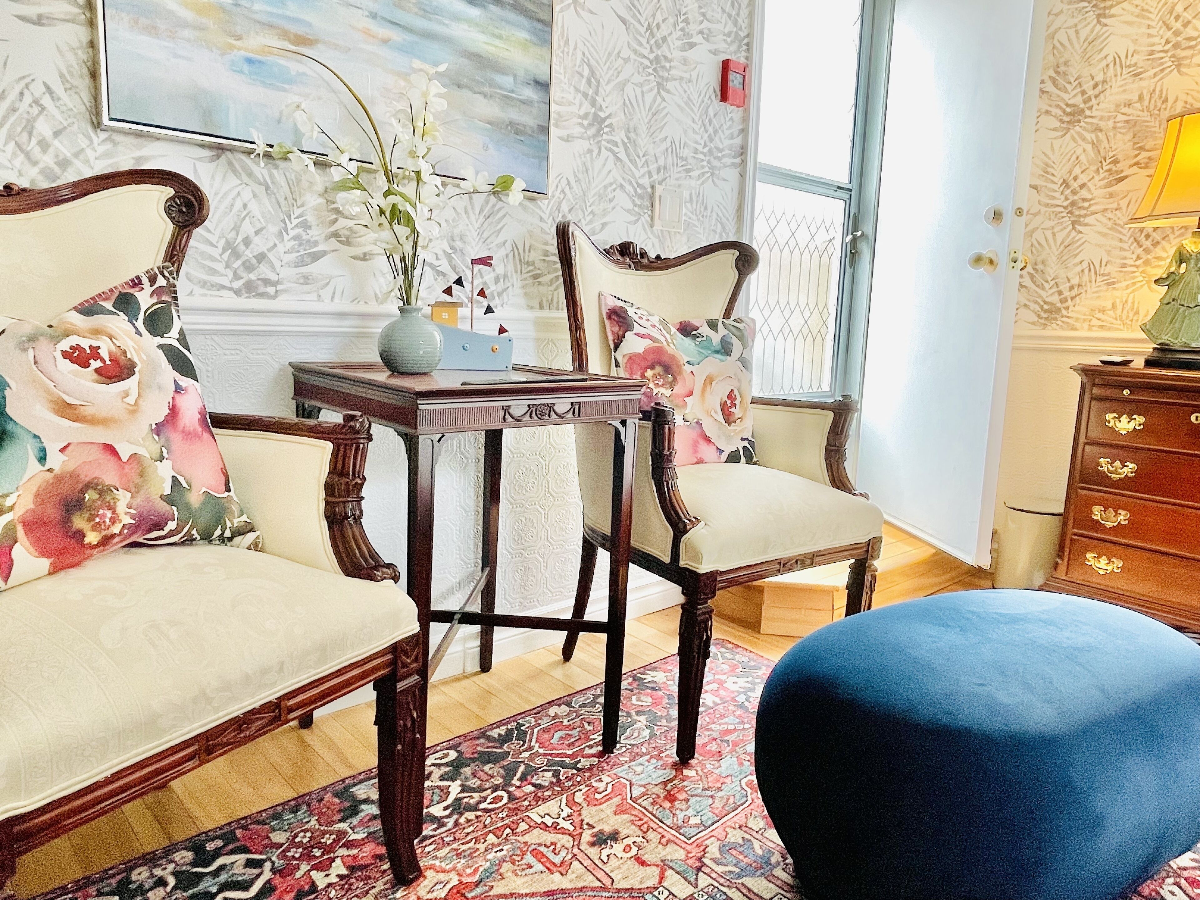 A room with two ornate wooden chairs, a small table with a vase and a book, a blue ottoman, and a patterned rug.