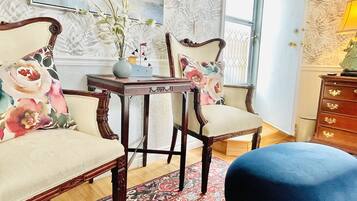 A room with two ornate wooden chairs, a small table with a vase and a book, a blue ottoman, and a patterned rug.