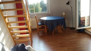 Huoneessa on puinen portaikko, pyöreä pöytä, jonka päällä on sininen pöytäliina, valkoinen sohva ja ikkuna, josta avautuu näkymä puihin.