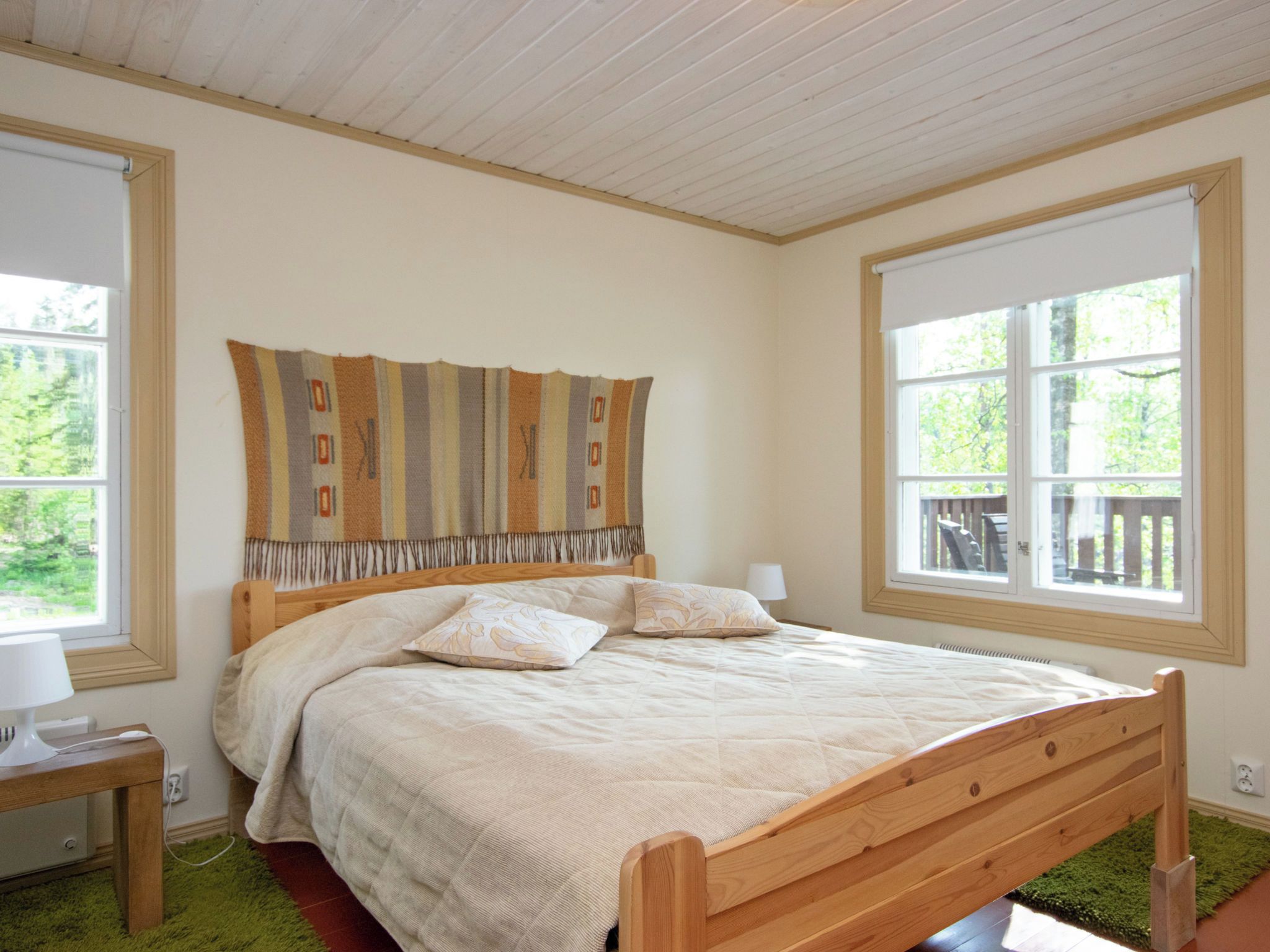 A bedroom with a wooden bed, a headboard with a patterned throw, two bedside lamps, and large windows with a view of greenery.