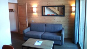 A room with a wooden wall, a gray sofa, a small table with a book, and a mirror reflecting a staircase. - Superb 6-person apartment at the foot of the slopes, in the "Coeur des Loges" residence (Les Menuires)