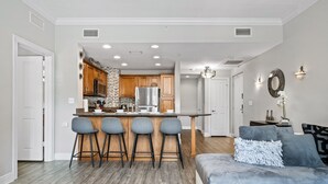 A modern kitchen with wooden cabinets, a central island with bar stools, and a dining area with a grey sofa and decorative pillows. - Beautiful Condo Near Amenities and Attractions (Kissimmee)