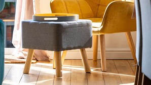 Una habitación con una silla amarilla, un reposapiés gris y una ventana con cortinas.