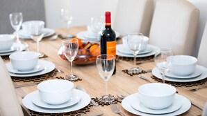 Una mesa de comedor servida con platos, tazones y copas de vino blancos, con una botella de vino y un bol de fruta en el centro.