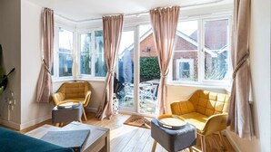 Una habitación luminosa con ventanas grandes, un sofá amarillo, una mesita y una silla azul.
