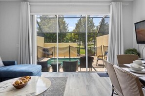 Relax in this spacious living room with a sofa, coffee table, and wicker chair, leading to an outdoor area with a pool and seating.