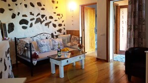 Un salon chaleureux avec un mur en pierre, un canapé agrémenté de coussins à motifs, une table basse en bois sur laquelle reposent une bouteille et des oranges, et un fauteuil noir.