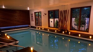 An indoor swimming pool with candles along the edge, wooden walls, and windows with views of trees.