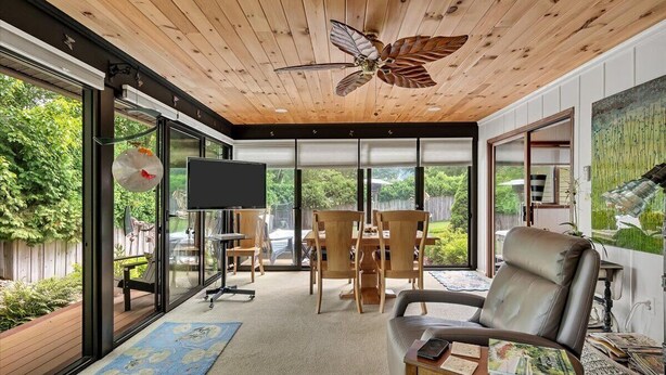 A modern living room with a wooden ceiling, a ceiling fan, a TV, a dining area with chairs and a table, and large windows offering a view of the outdoors.
