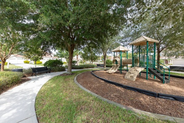Explore a shaded playground with a slide and climbing structure, surrounded by mature trees and benches along a winding path.