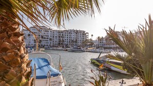 Un puerto deportivo con barcos amarrados, un edificio de varios pisos al fondo y una palmera en primer plano.