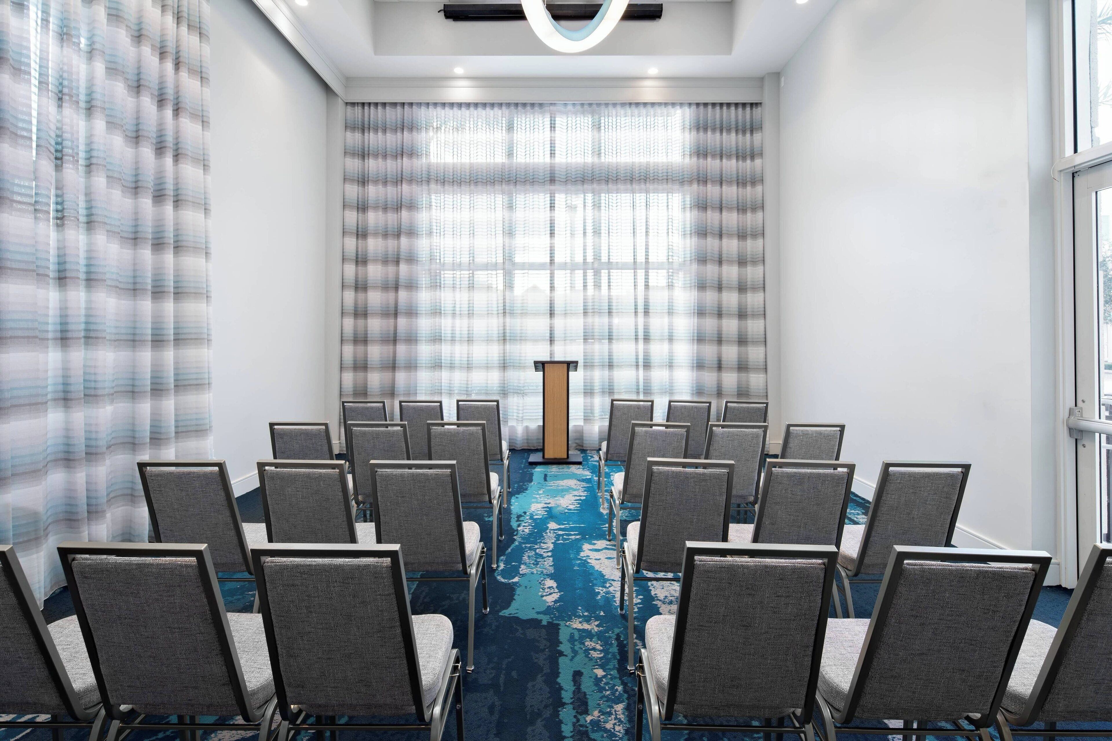 A conference room with rows of chairs facing a podium, a blue carpet, and large windows with curtains.
