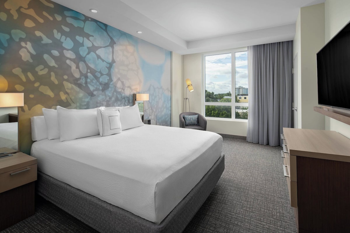A hotel room with a large bed, a desk, a chair, a TV, and a window with curtains.