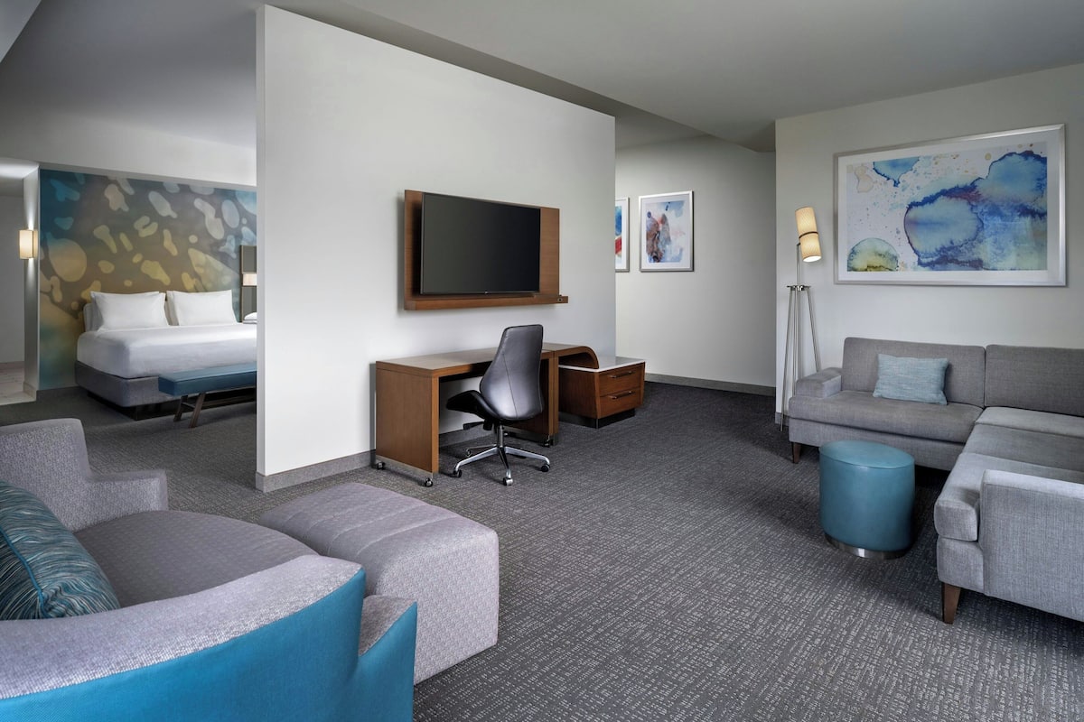 A hotel room with a bed, a desk with a chair, a sofa, a blue ottoman, and a TV mounted on the wall.
