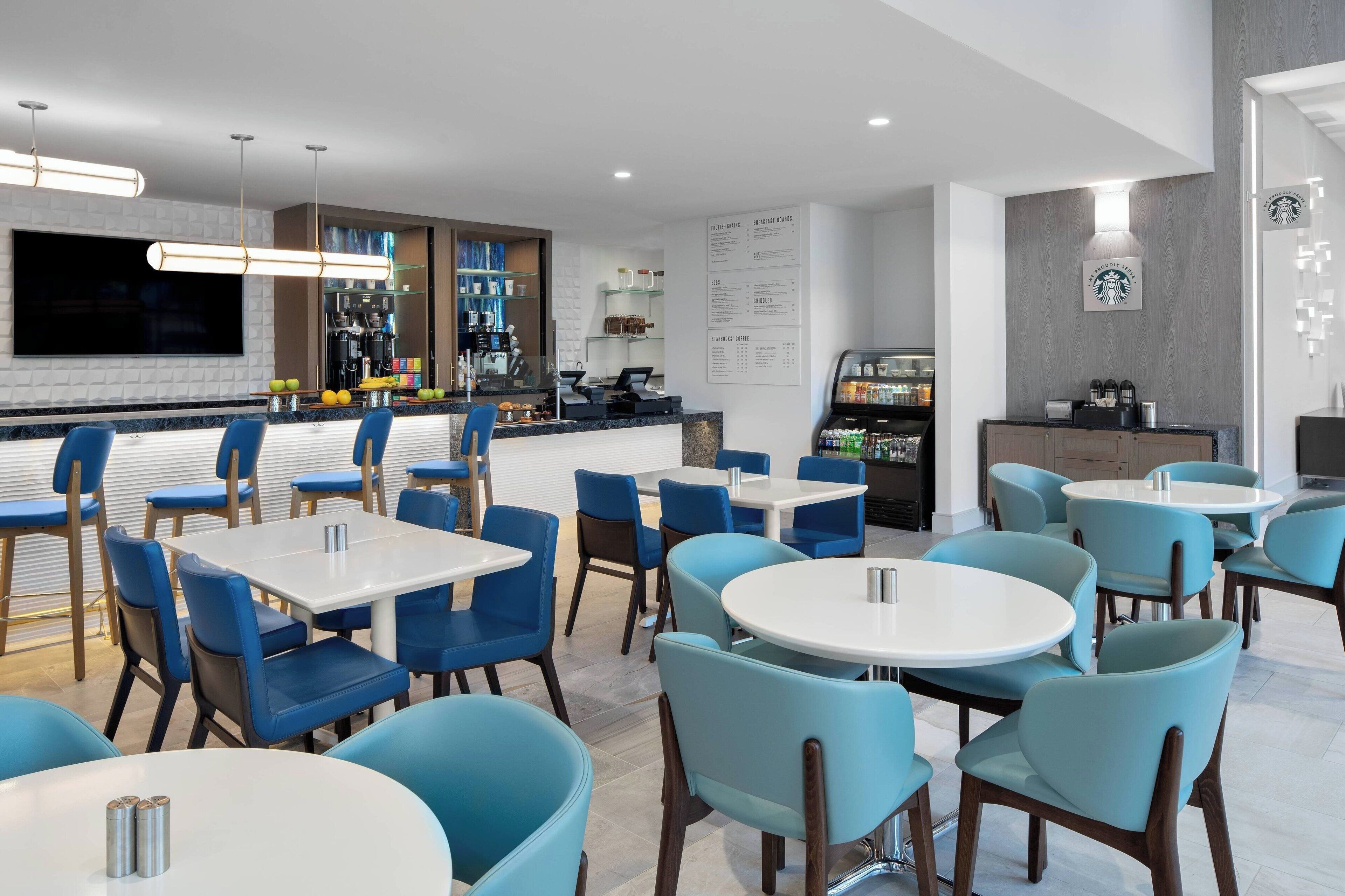 Un restaurante moderno con sillas azules y mesas blancas, una zona de bar y un panel con la carta.