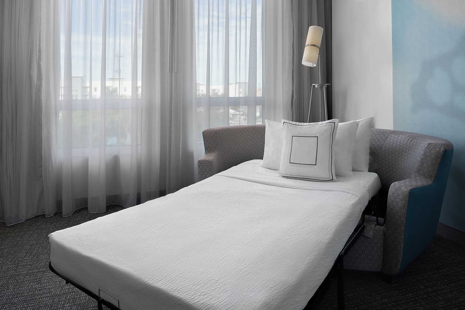 A hotel room with a single bed, a chair, a lamp, and sheer curtains.
