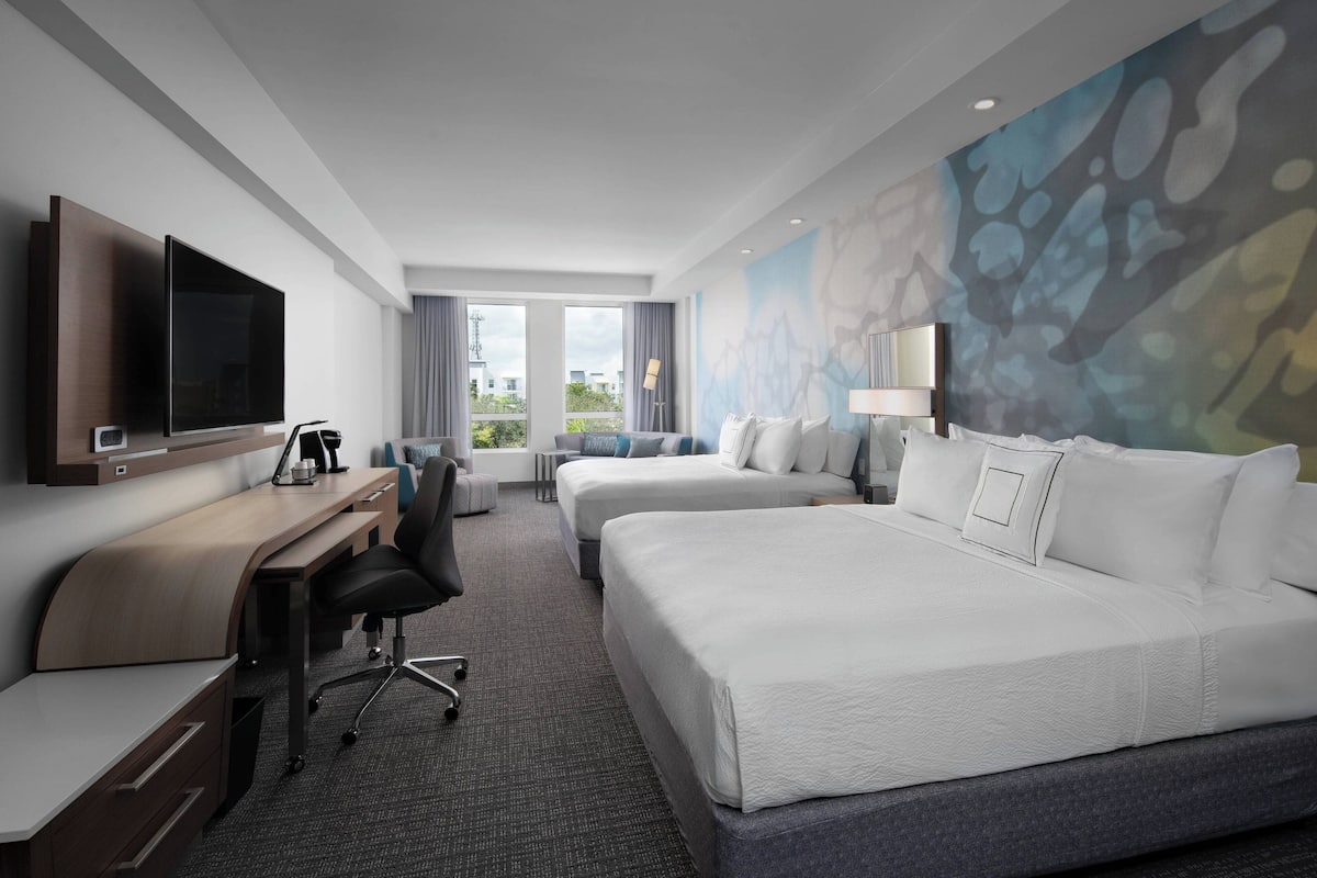 A hotel room with a flat-screen TV, a desk with a chair, two beds, and a large window with a view of buildings.