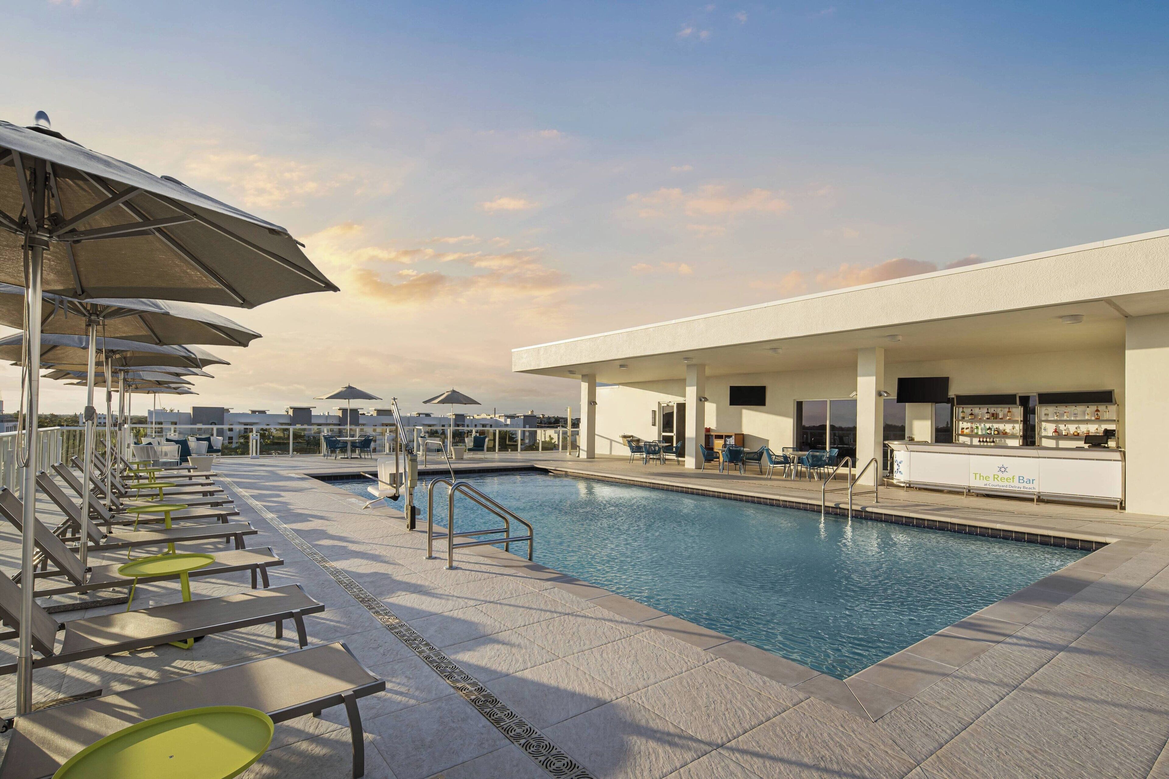 A rooftop pool area with sun loungers and umbrellas, a bar, and a pool with a view of the city skyline.
