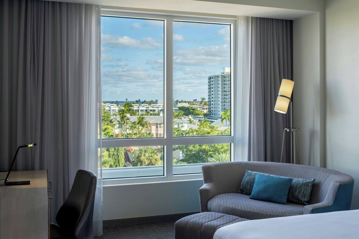 A hotel room with a large window, a desk, a sofa, and a floor lamp.
