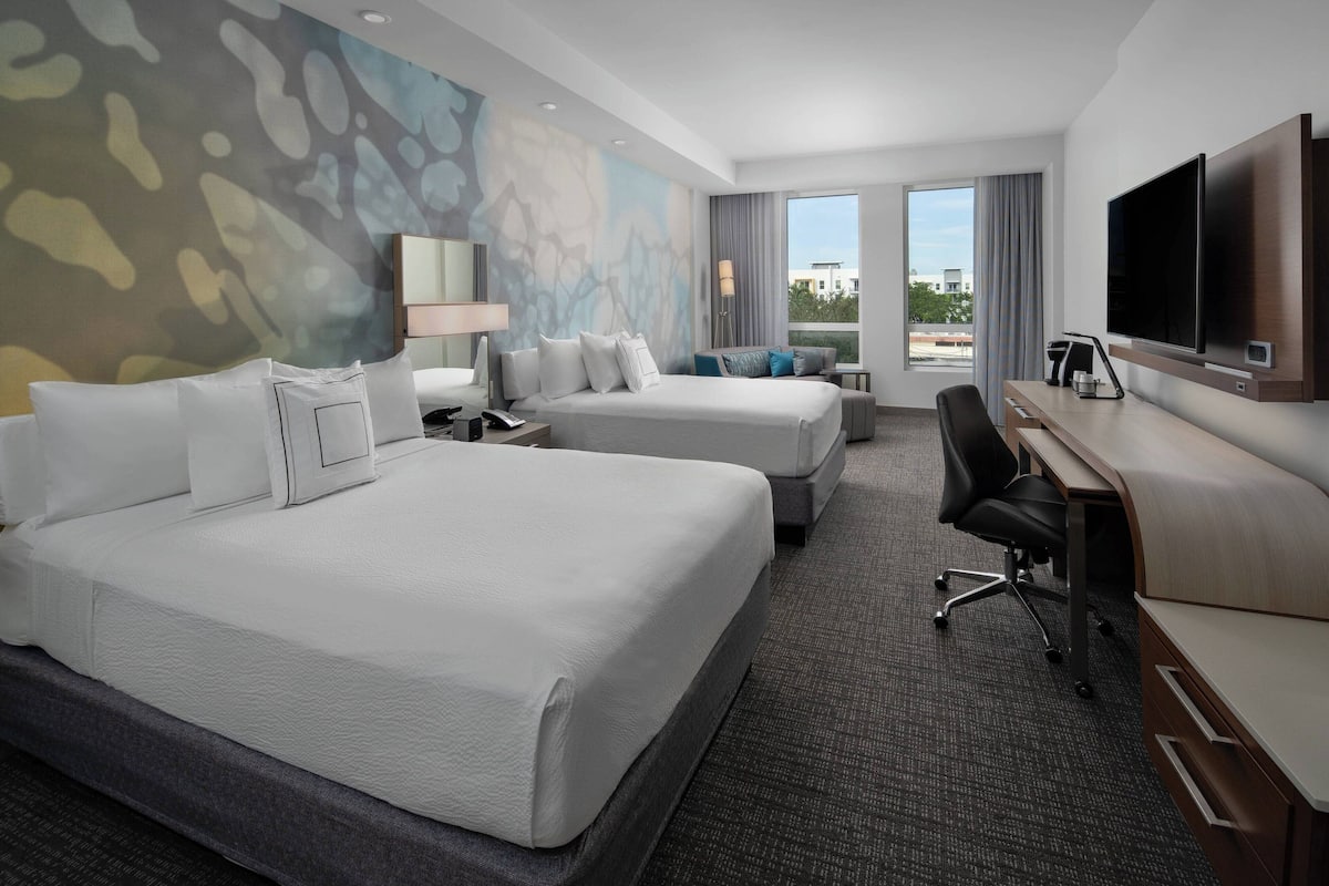 A hotel room with two beds, a desk, a TV, and a large window with a view.