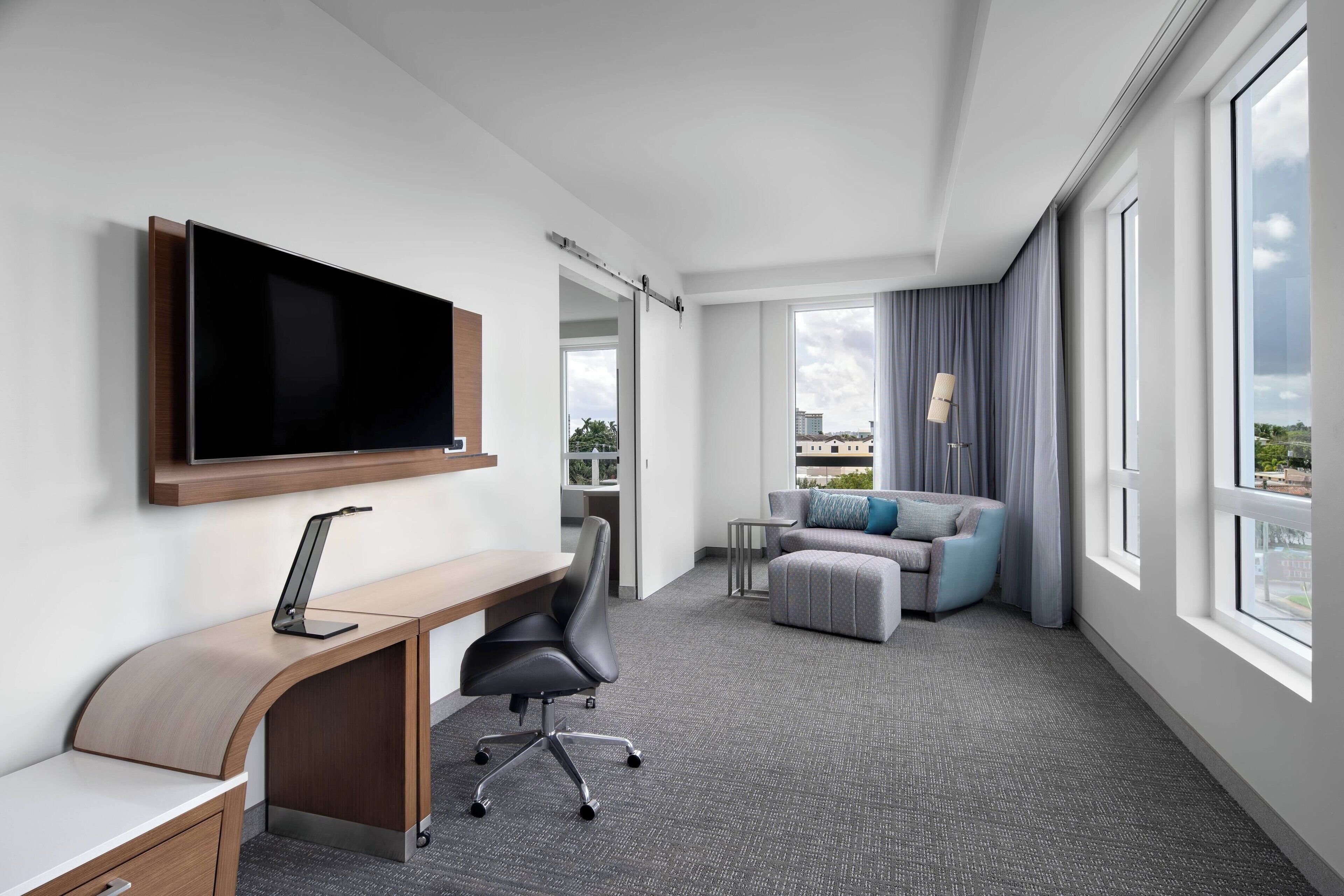 A modern hotel room with a flat-screen TV mounted on the wall, a desk with a laptop, a comfortable sofa, and large windows offering a city view.