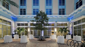 A hotel courtyard with a central tree, surrounded by tables and benches, and large windows on the building facade. - Courtyard by Marriott Delray Beach (Delray Beach)