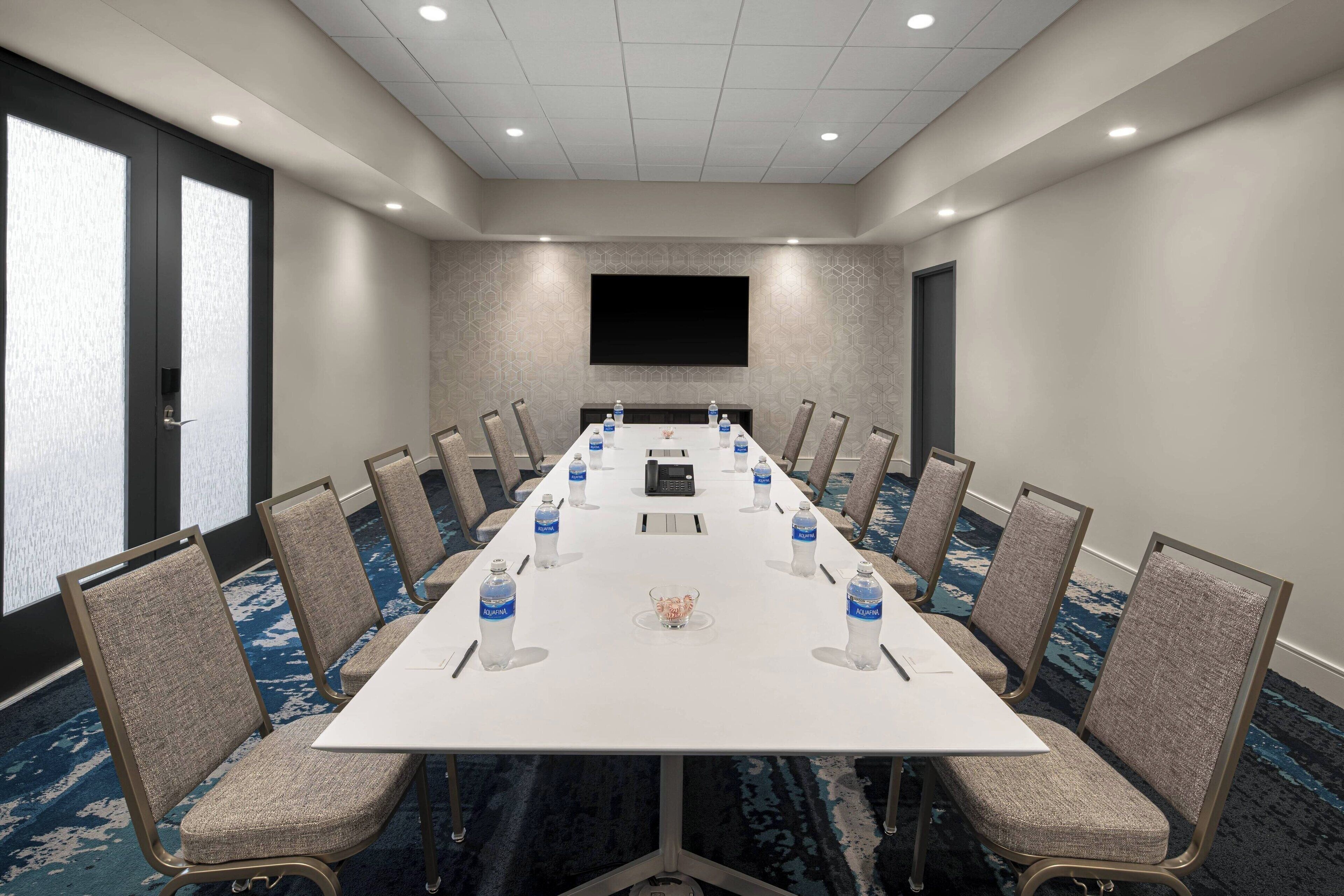A conference room with a long table, chairs, water bottles, a small decorative item, and a TV screen on the wall.