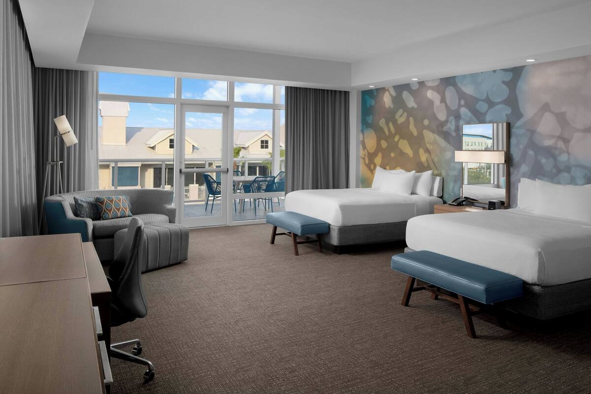A hotel room with two beds, a desk, a chair, and a large window offering a view of the outside.