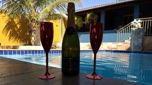 Una botella de champán y dos copas junto a una piscina, con una palmera y un edificio al fondo.