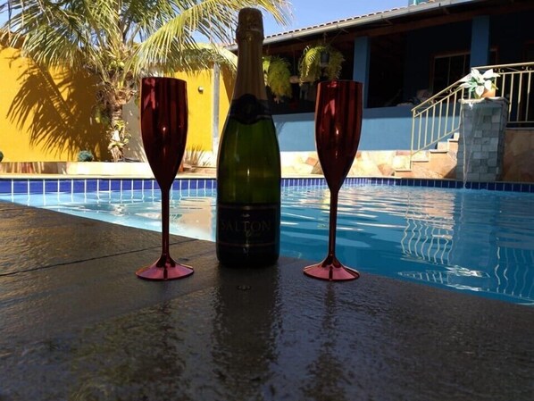 A bottle of champagne and two glasses on a poolside with a palm tree and a building in the background.