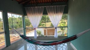 Una hamaca colgada entre dos ventanas con cortinas translúcidas, un suelo reflectante y vistas al verde exterior.