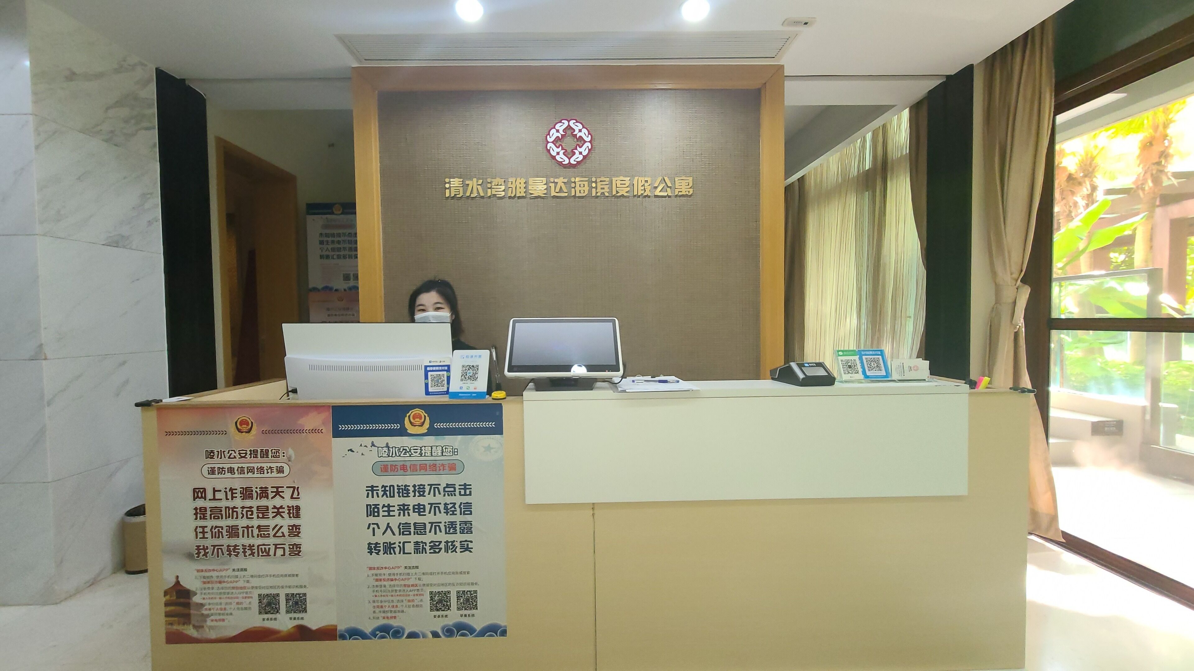 A reception desk with a computer, a monitor, and a QR code scanner. There are posters on the wall and a window with curtains.
