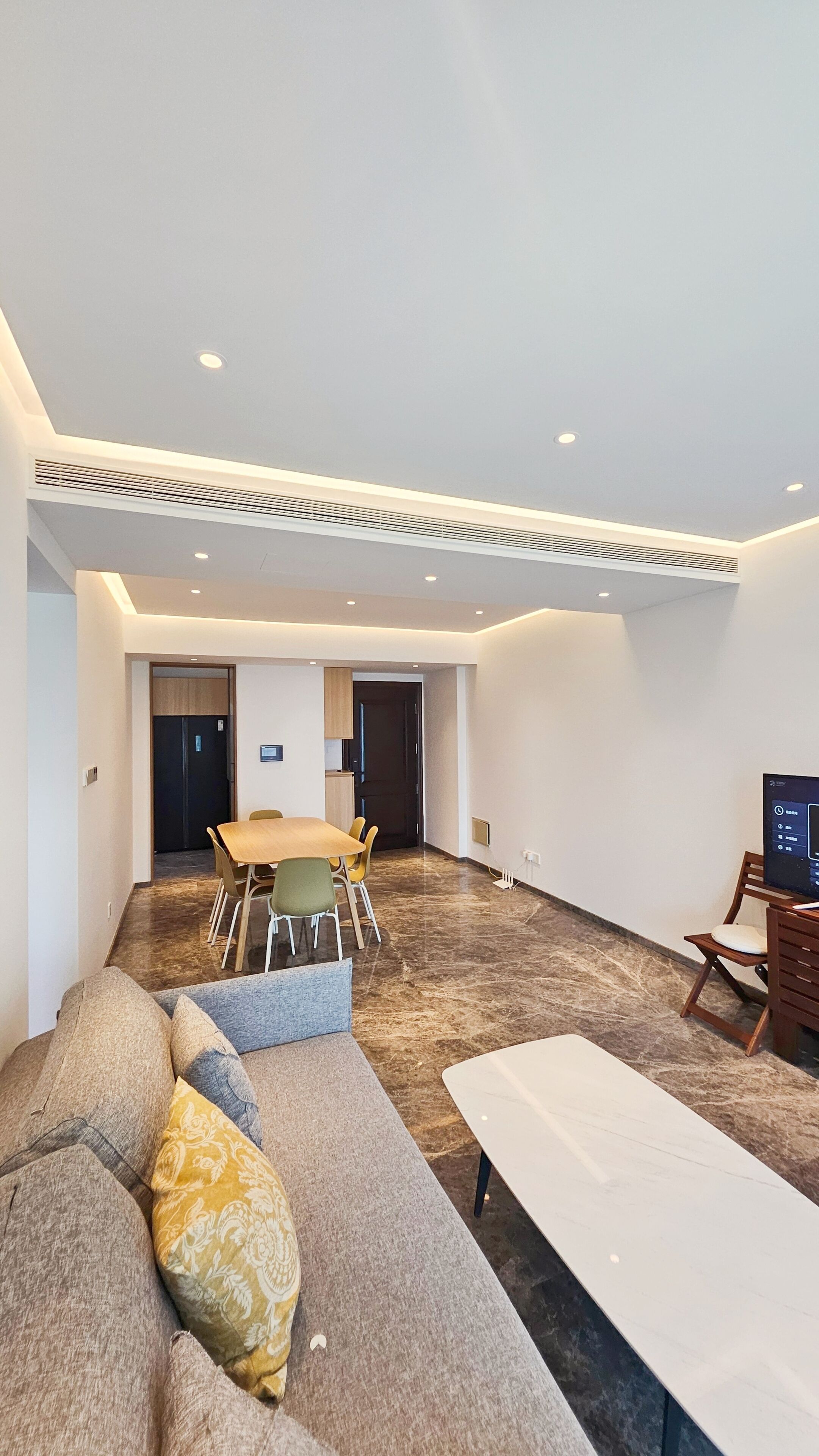 A modern hotel room with a sofa, a dining table with chairs, a TV, and a large window.