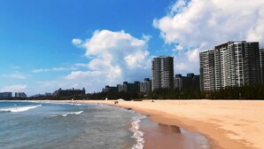 A beach with a sandy shore and ocean waves, with tall buildings in the background. - Amanda Seaview Resort Apartment (Lingshui)